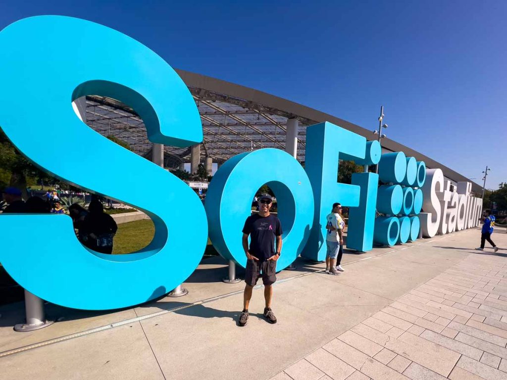 Dan Brewer, of UltimateSportsRoadTrip.com, stands in front of the SoFi Stadium sign before a Los Angeles Rams home game.