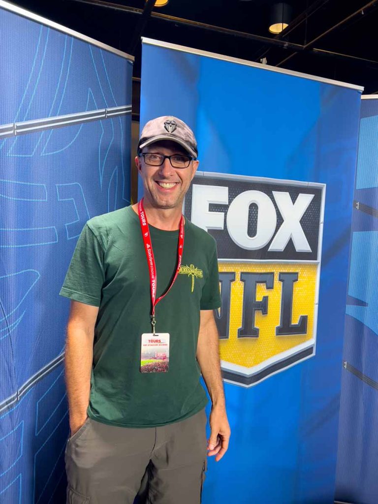 Dan Brewer, owner of UltimateSportsRoadTrip.com, stands in front of a FOX NFL sign in the broadcast booth during a State Farm Stadium tour the day before an Arizona Cardinals game.